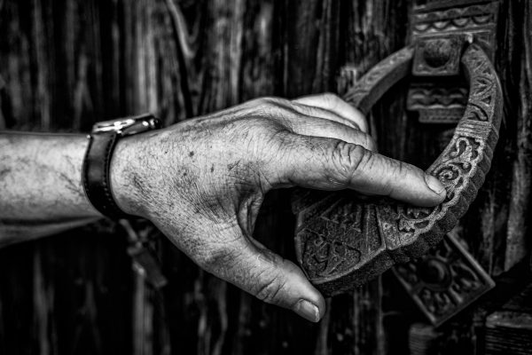 Detailed black and white photo of a hand gripping an intricately designed door knocker on a wooden gate